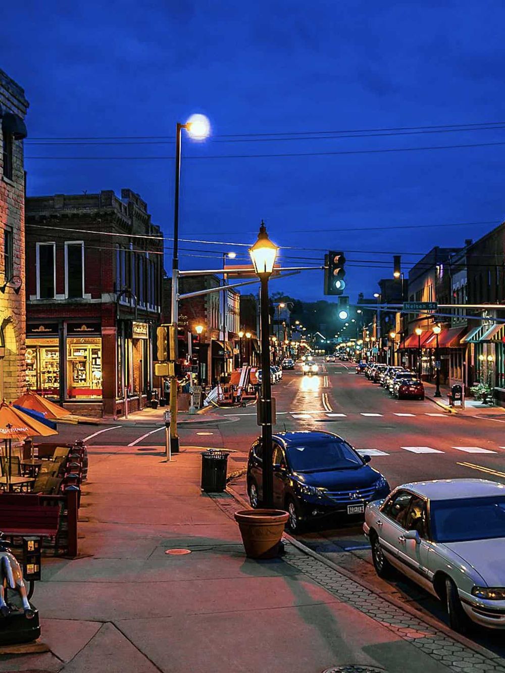 Night street scene with illuminated storefronts and parked cars, vibrant city nightlife atmosphere.