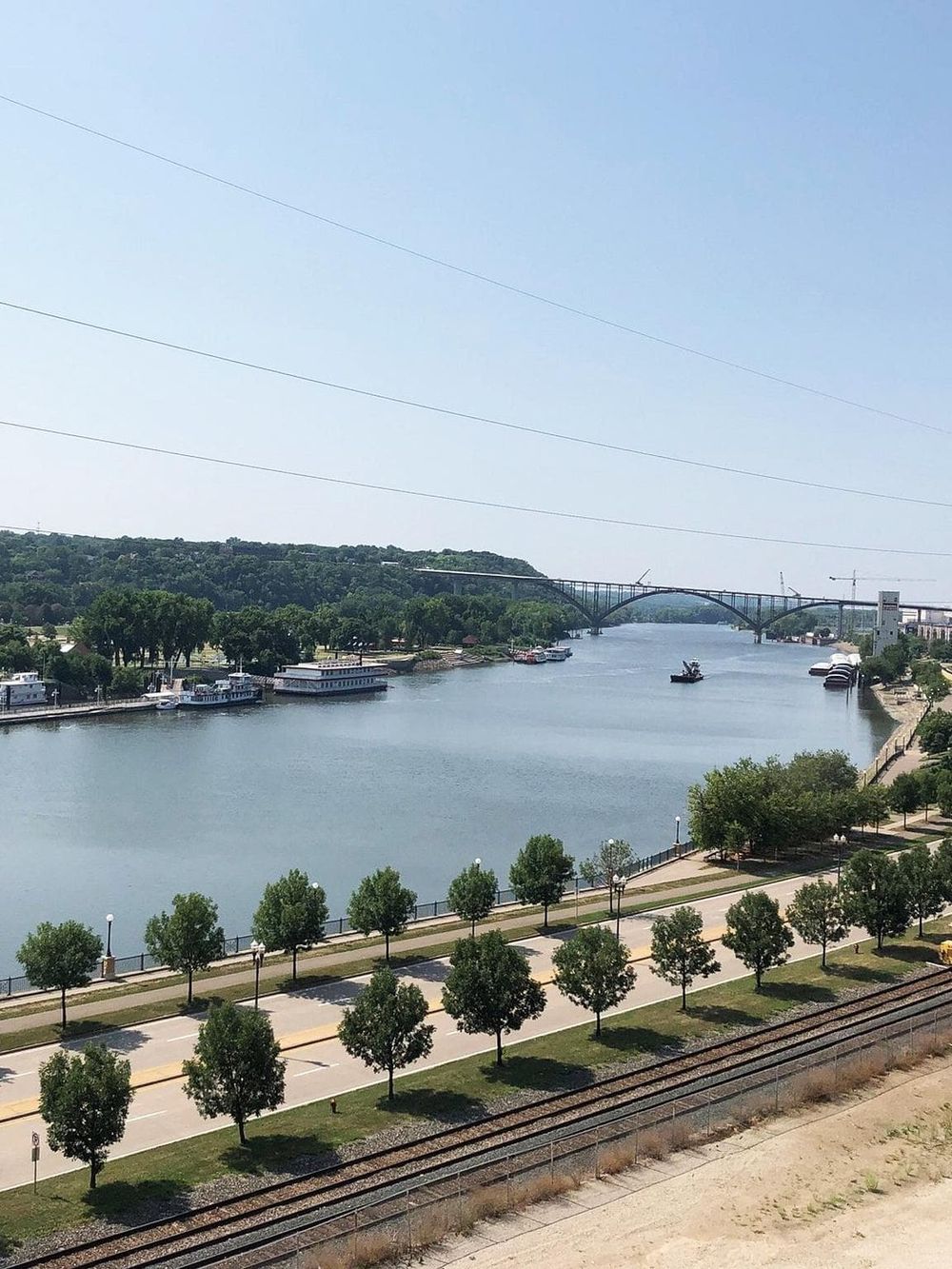 Bright cityscape with river, bridge, boats, and waterfront park, showcasing scenic views and urban waterways.