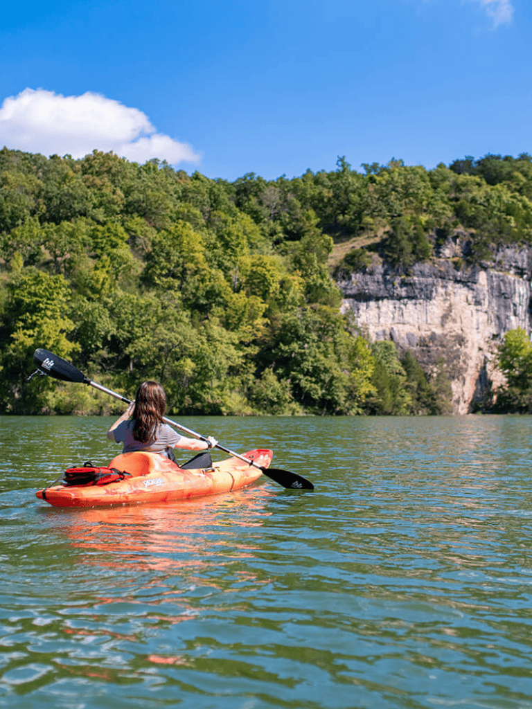 Kayaking on a serene river surrounded by lush green trees and rocky cliffs under a bright blue sky.