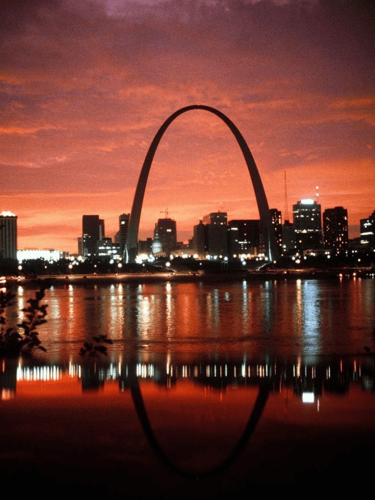 St. Louis skyline with Gateway Arch during sunset, reflecting on the river, city lights illuminating the scene.