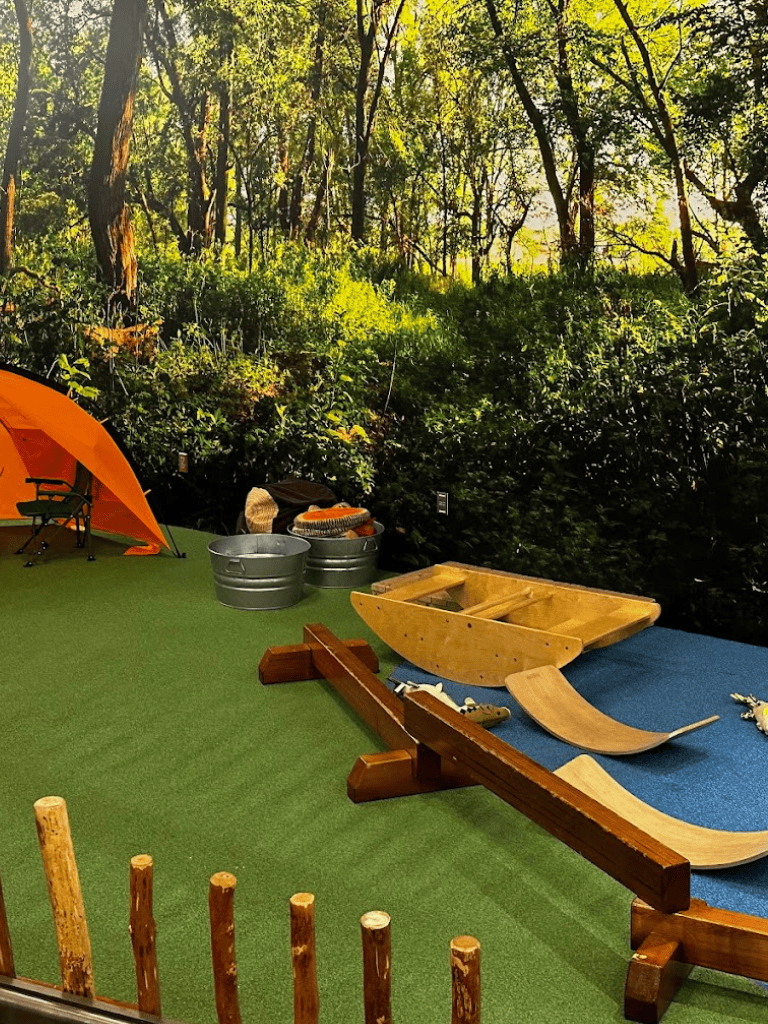 Bright outdoor camping setup with wooden boats, orange tent, and green forest background.