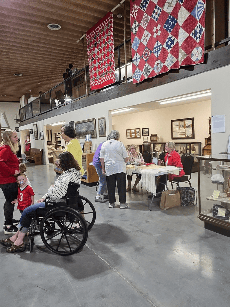 Handicapped woman at craft fair with multiple shoppers and quilt display in the background.
