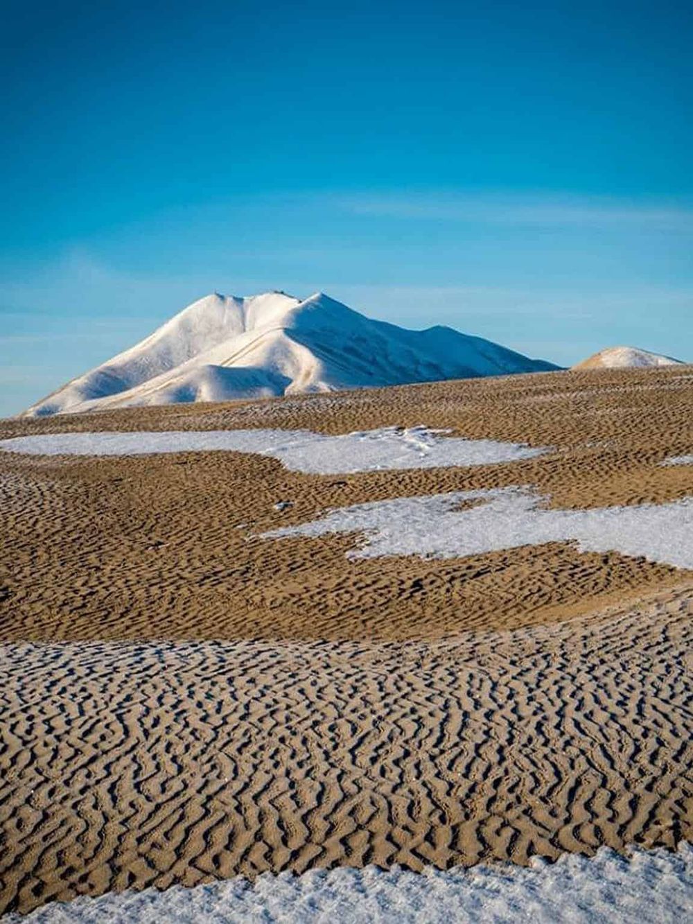 Snow-capped mountain with sandy desert landscape under clear blue sky.