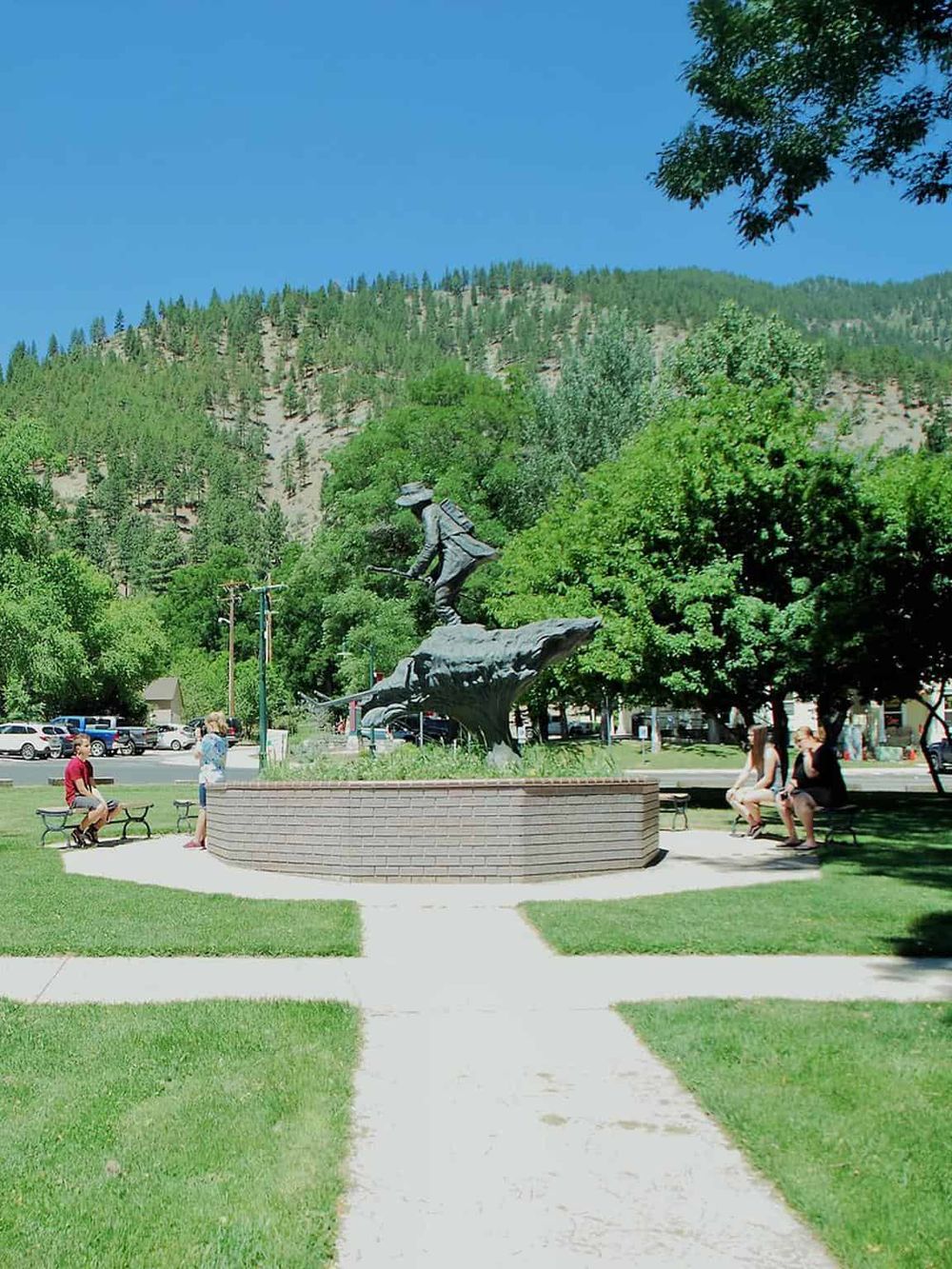 Bronze cowboy statue in park surrounded by trees, mountains, and visitors, symbolizing the quest for outdoor adventure.