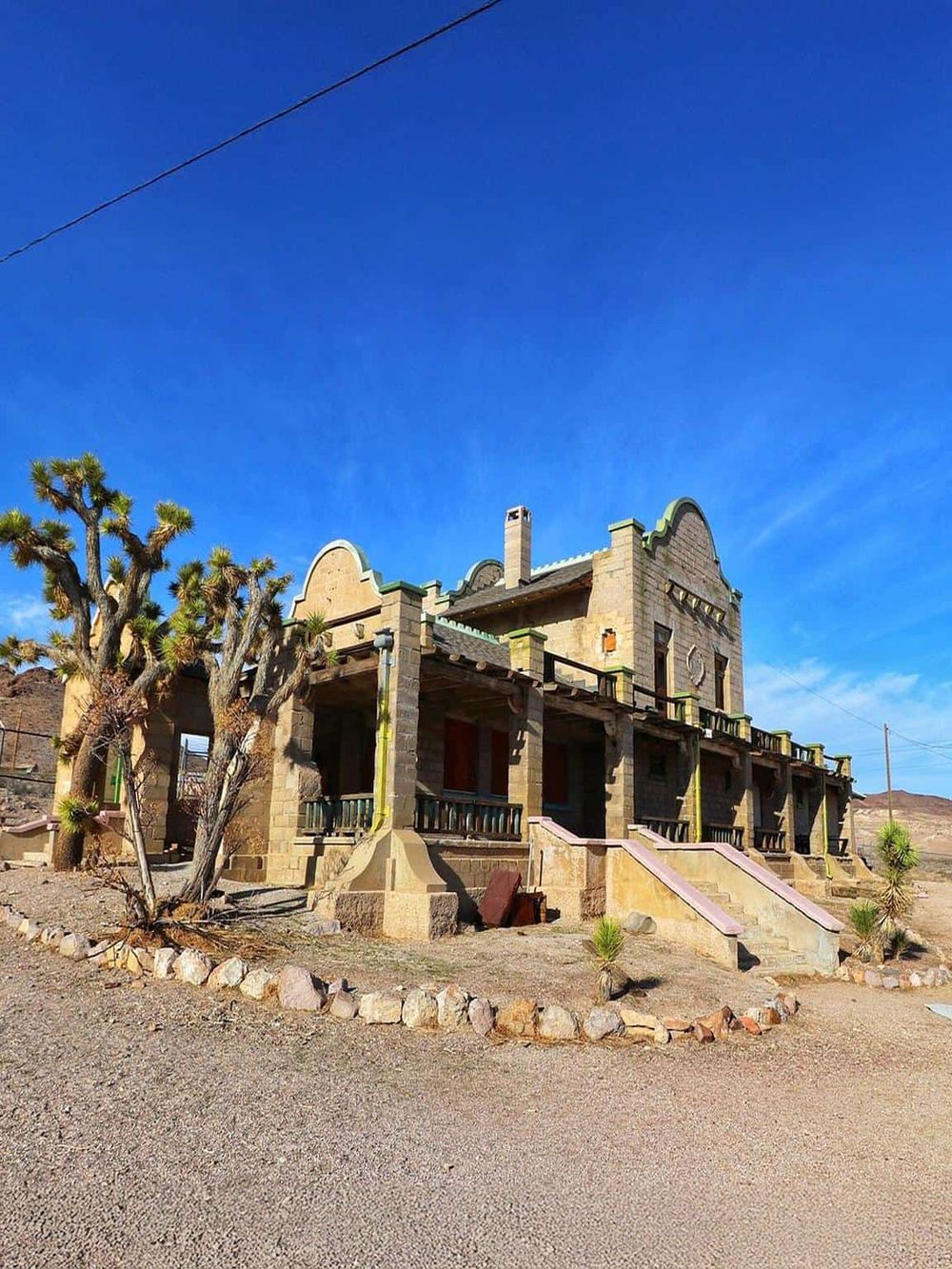 Old Western-style building in desert landscape with Joshua trees, clear blue sky, and rugged terrain.