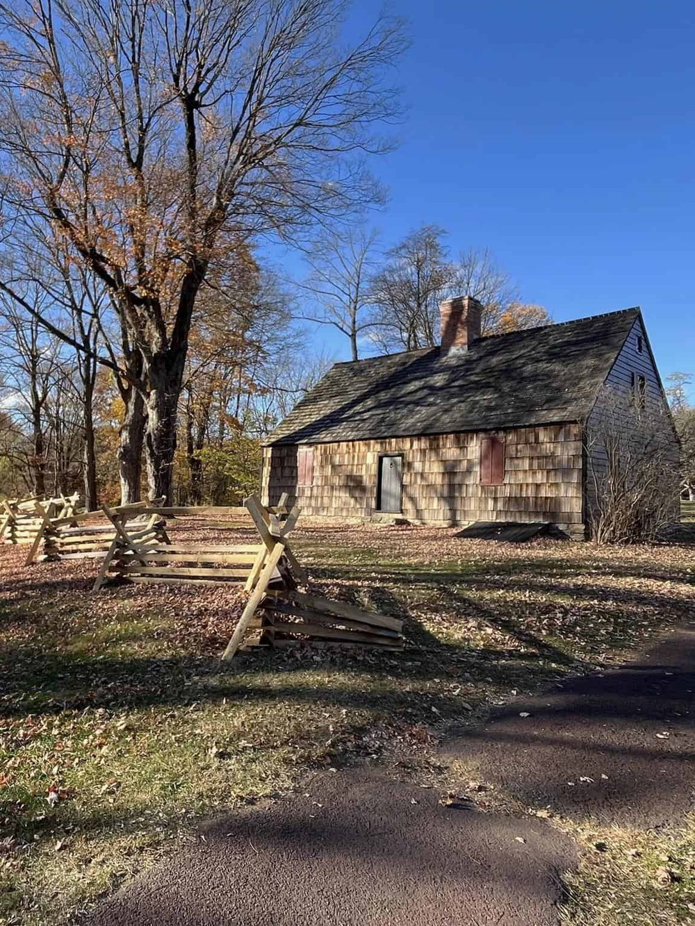Historic wooden house with leafless trees and fallen autumn leaves on the ground, set against a clear blue sky.