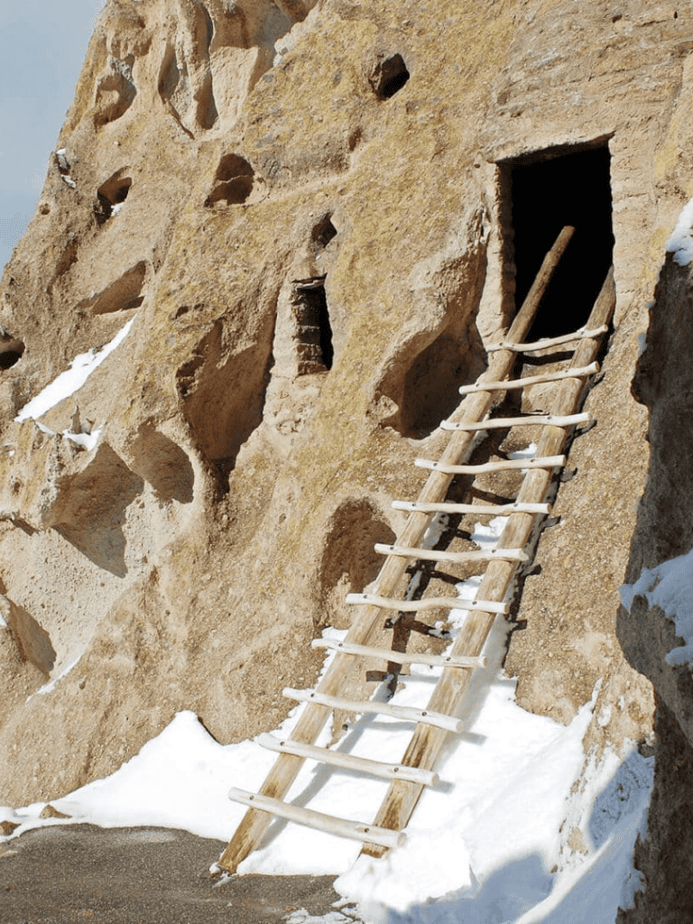 Cliffside ancient dwelling with wooden ladder and snow, archaeological site exploration in winter.