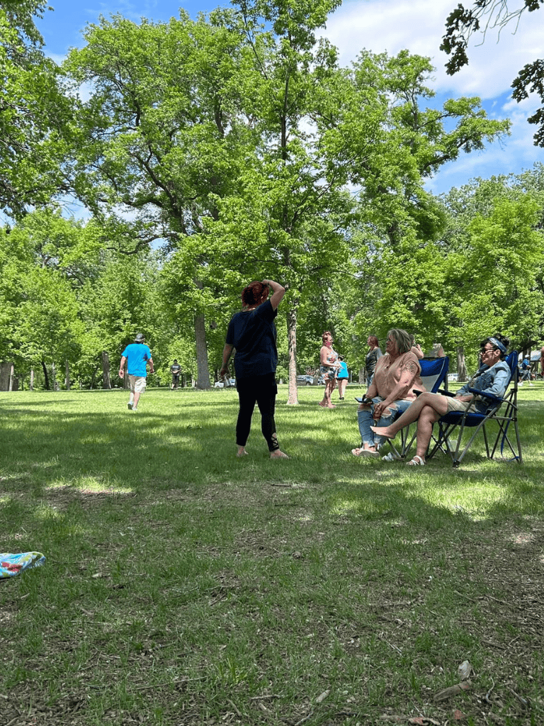 Relaxed outdoor park scene with people enjoying sunny day under lush green trees, sitting and walking peacefully.