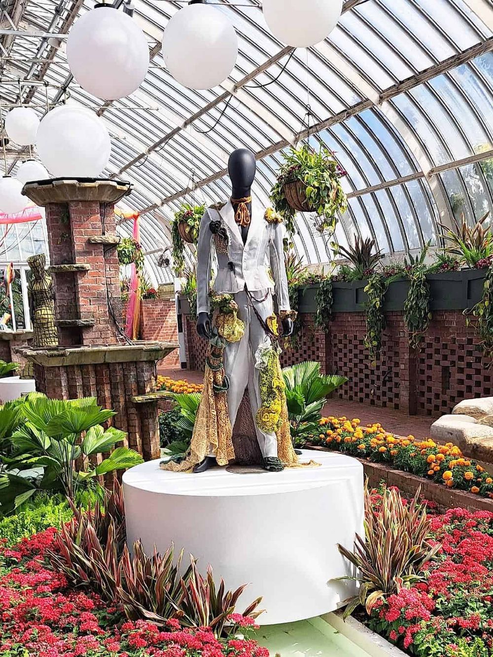 Colorful mannequin dressed in eclectic fashion inside a vibrant greenhouse filled with plants and flowers.