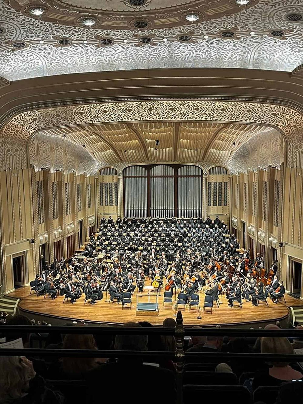 Orchestra performing in elegant concert hall with ornate ceiling and wooden accents.