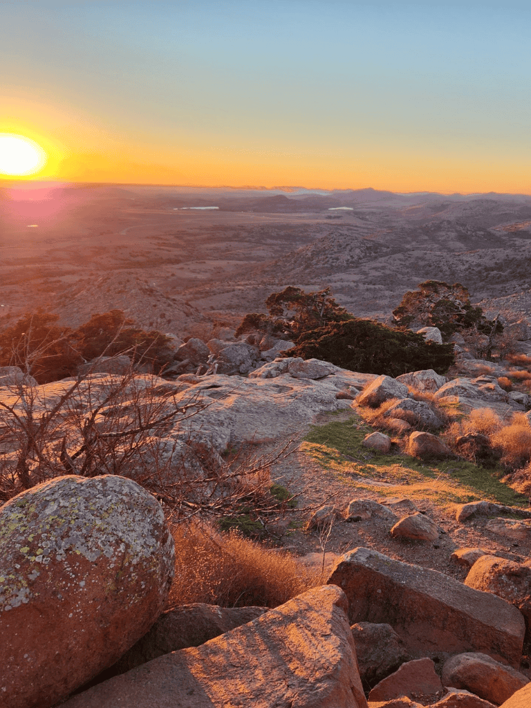 Vast rocky desert landscape during sunset with orange and pink hues, rugged terrain, and sparse vegetation.