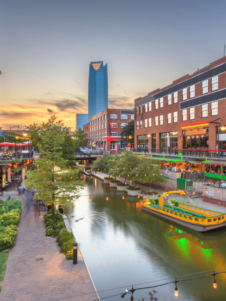 Vibrant downtown district with riverwalk, modern skyscraper, shops, restaurants, and evening lights.