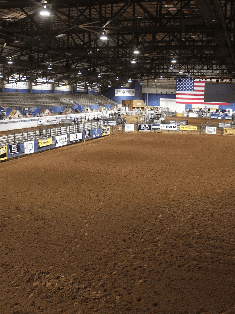 Arena du arena rodeo stadium with American flag, seating, and sponsorship banners for events and rodeo competitions.