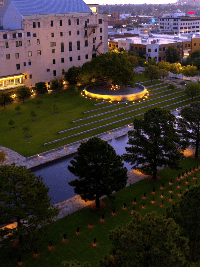 Modern city park with illuminated circular fountain, lush trees, and landscaped lawn at dusk.