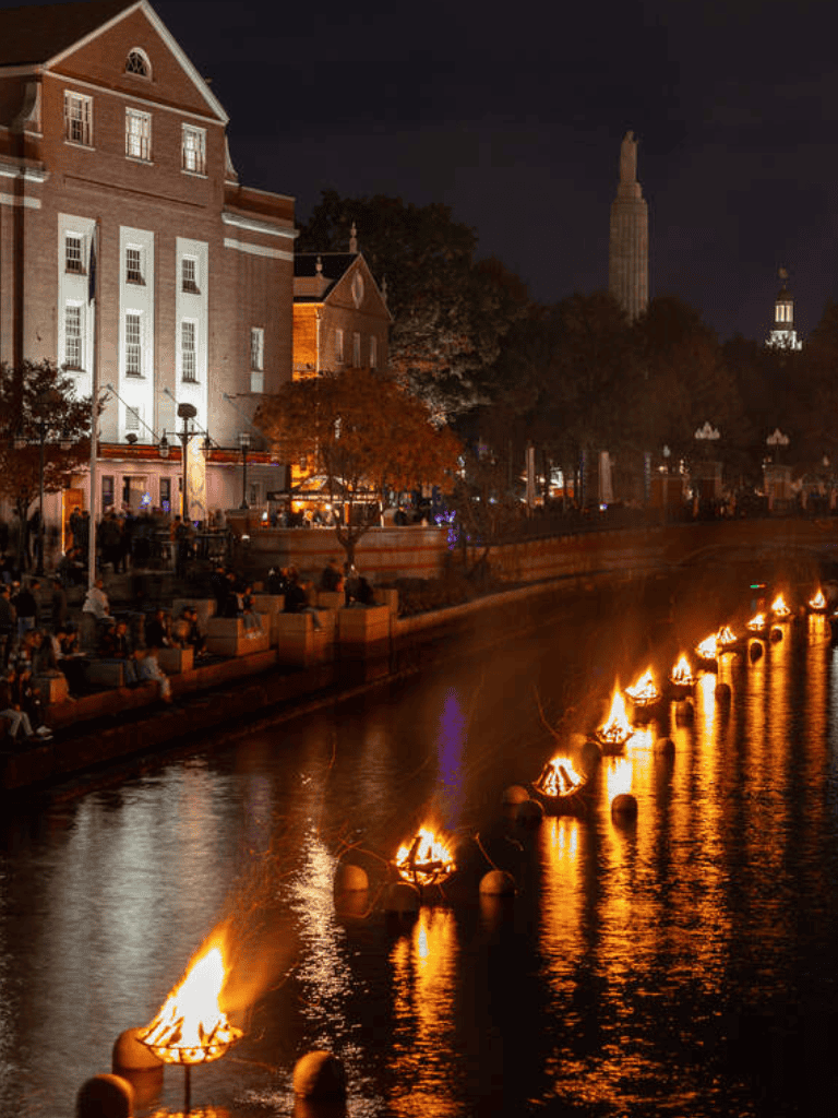 Nighttime cityscape with lit riverside fire pits, historic buildings, and iconic skyscraper skyline views in New York City.