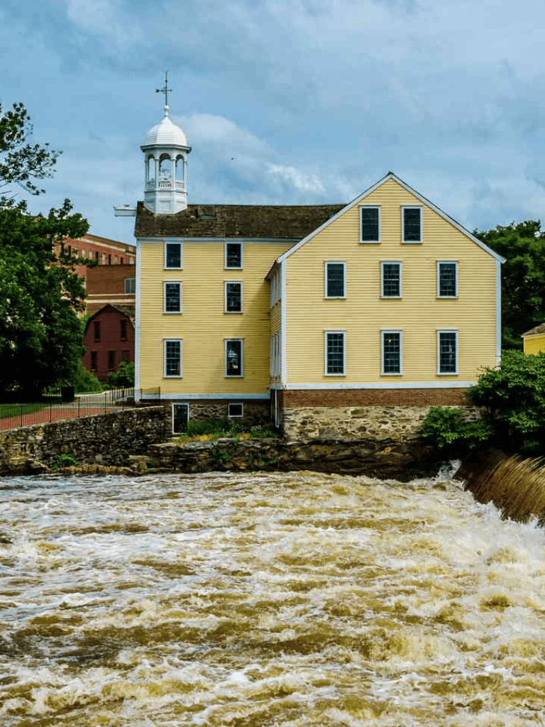 Historic yellow mill house on riverbank with flowing water and cloudy sky, in QuestForDirections location.