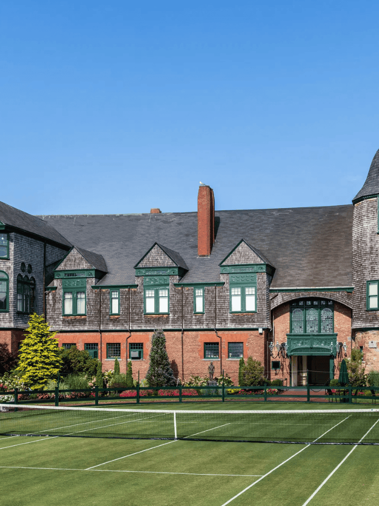 Victorian-style mansion with tennis court, lush landscaping, and bright blue sky.