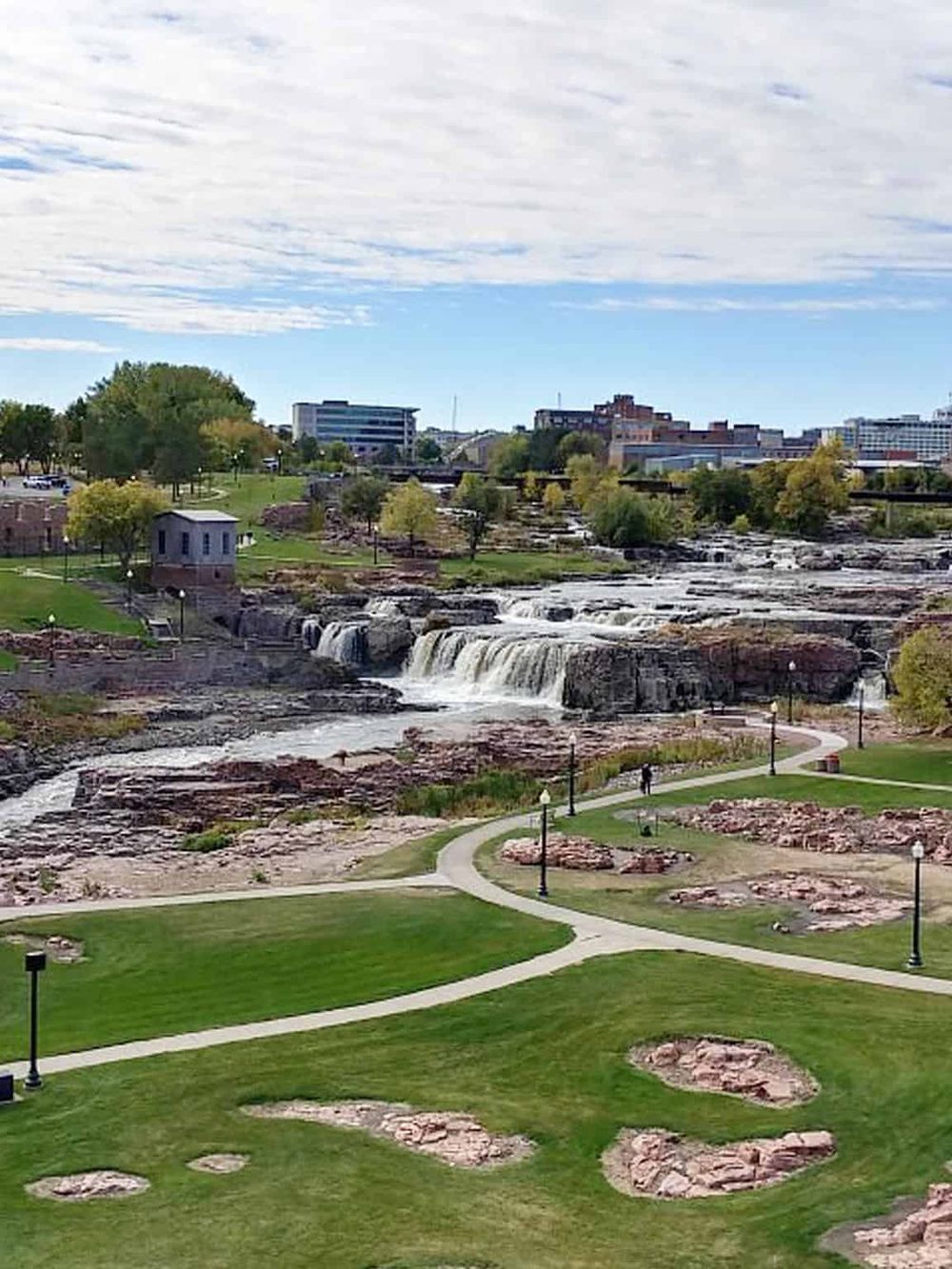 Serene park with waterfall, walking paths, lush greenery, and city skyline in the background. Perfect for outdoor recreation and relaxation.