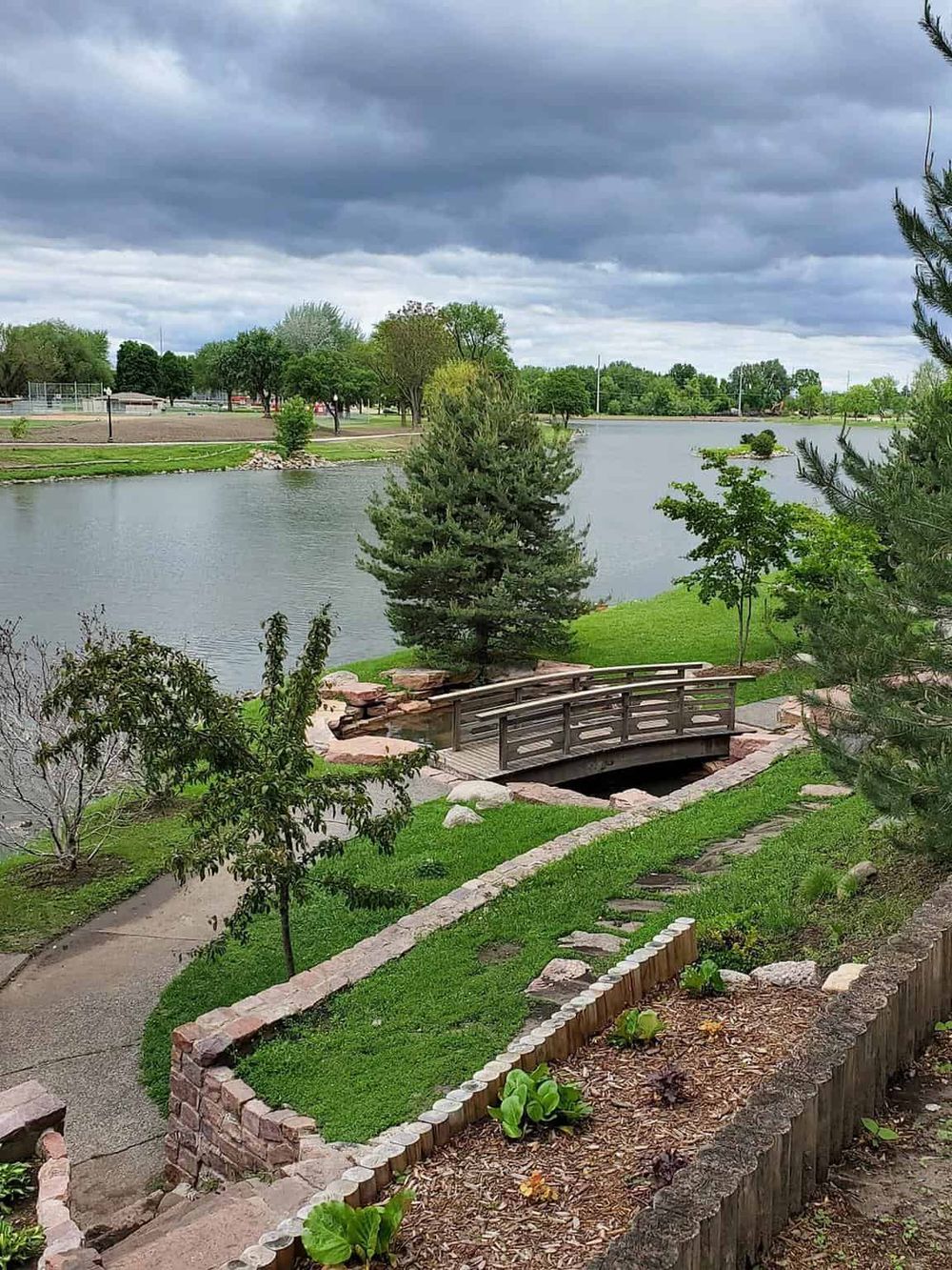 Serene park with a bridge over a pond, lush greenery, and dark stormy skies in a scenic outdoor setting.