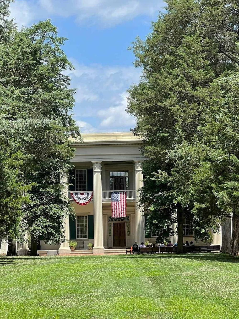 Historic mansion with large columns and American flags, surrounded by green trees and lawn, representing event venue or historical site.