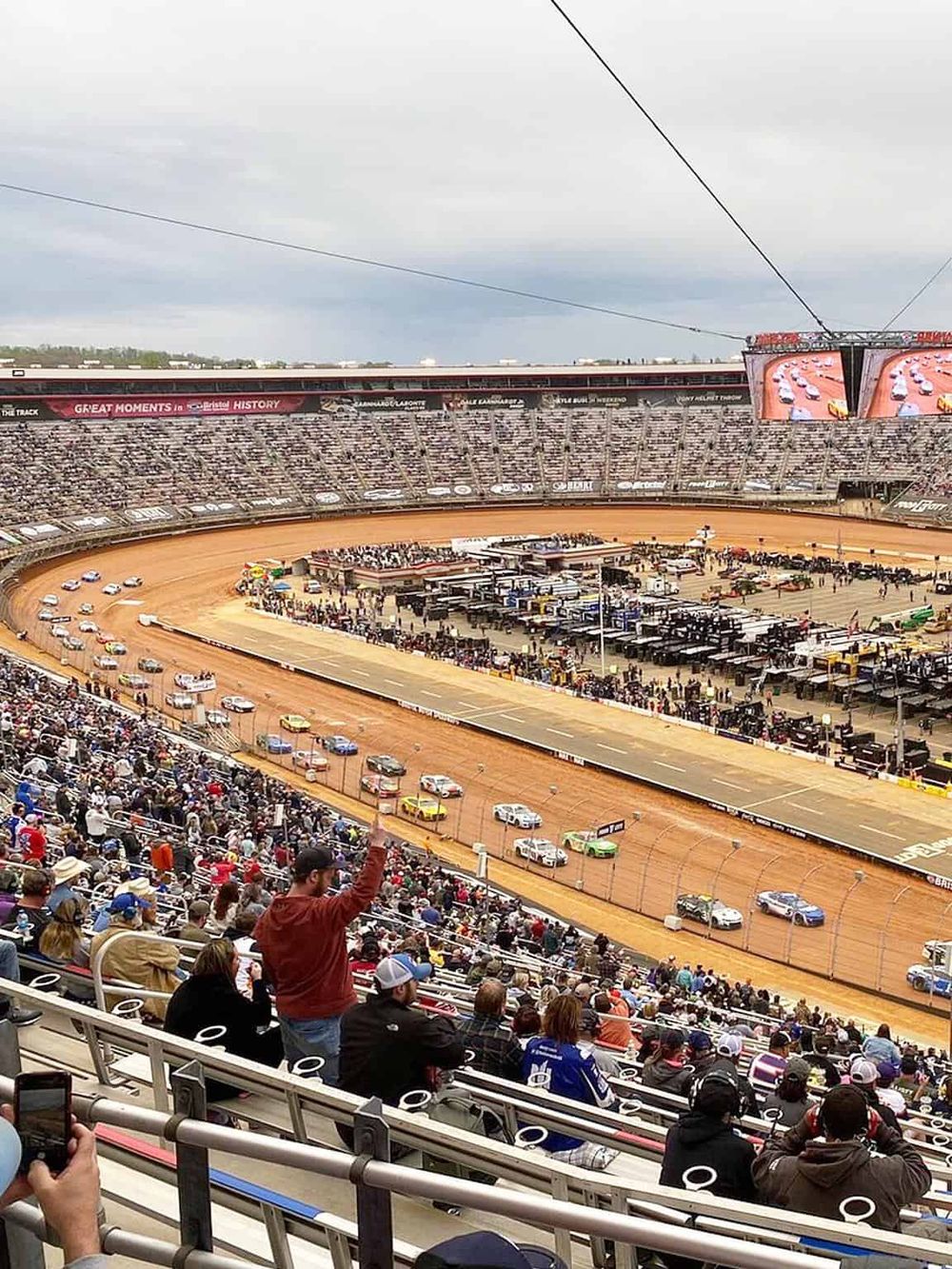 Cars racing on dirt track at a NASCAR race, large crowd watching from stands.