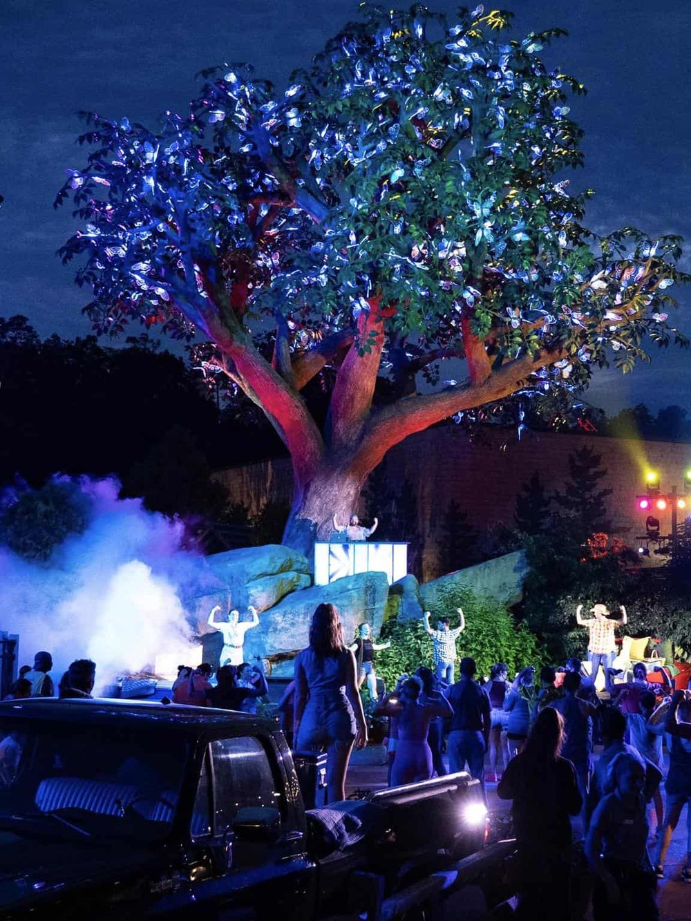 Colorful outdoor concert with lit-up tree and crowd at night, vibrant atmosphere, entertainment, music event, community gathering.