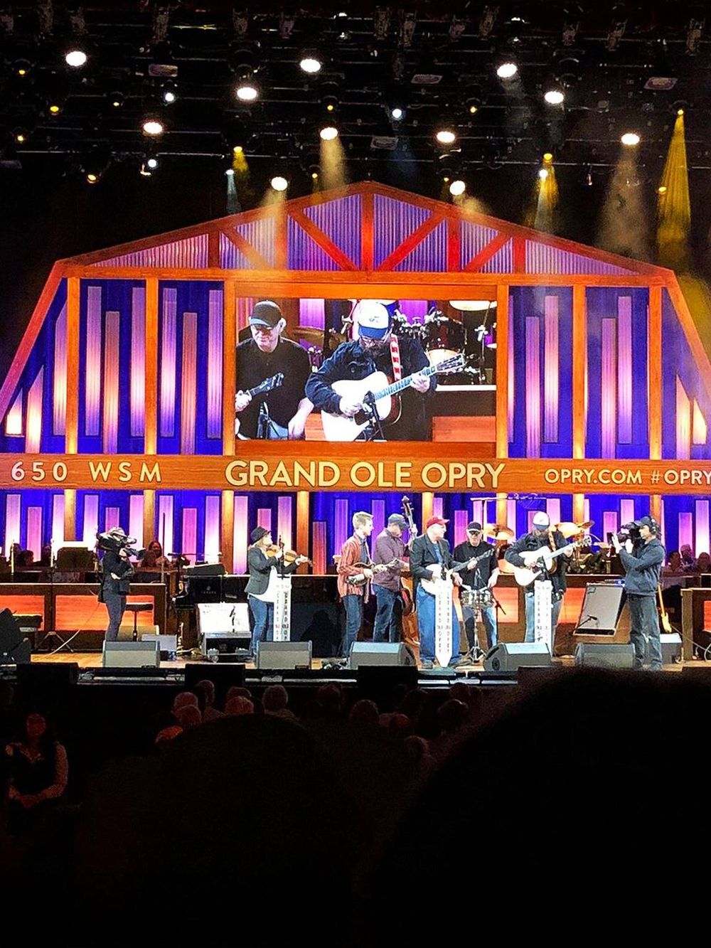 Vibrant concert stage at Grand Ole Opry featuring live musicians, colorful lighting, and a large video screen.