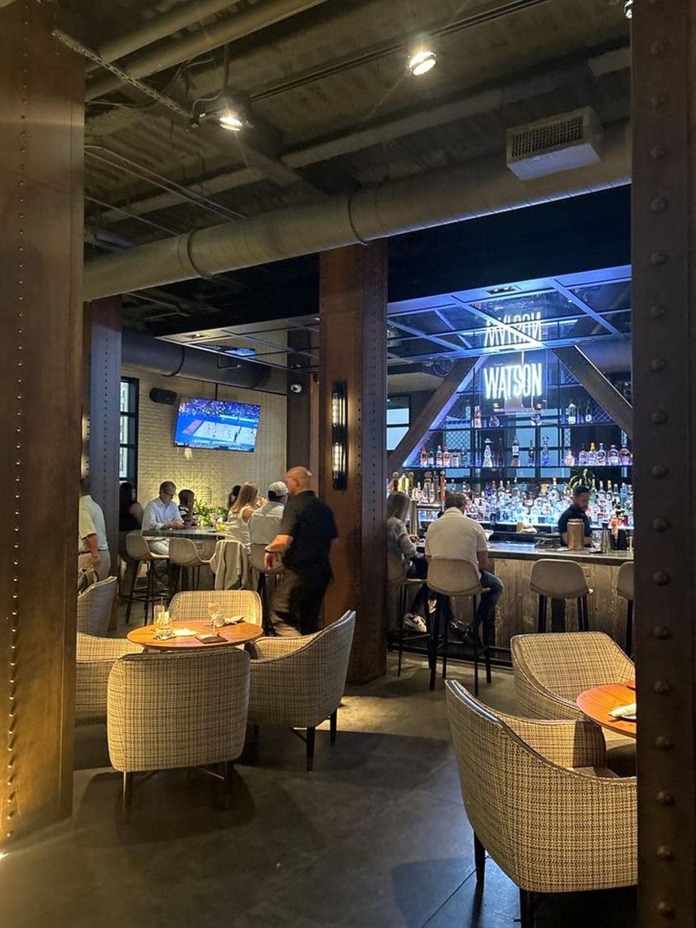 Cozy bar and restaurant interior with modern industrial decor, illuminated bar area, and patrons enjoying drinks.