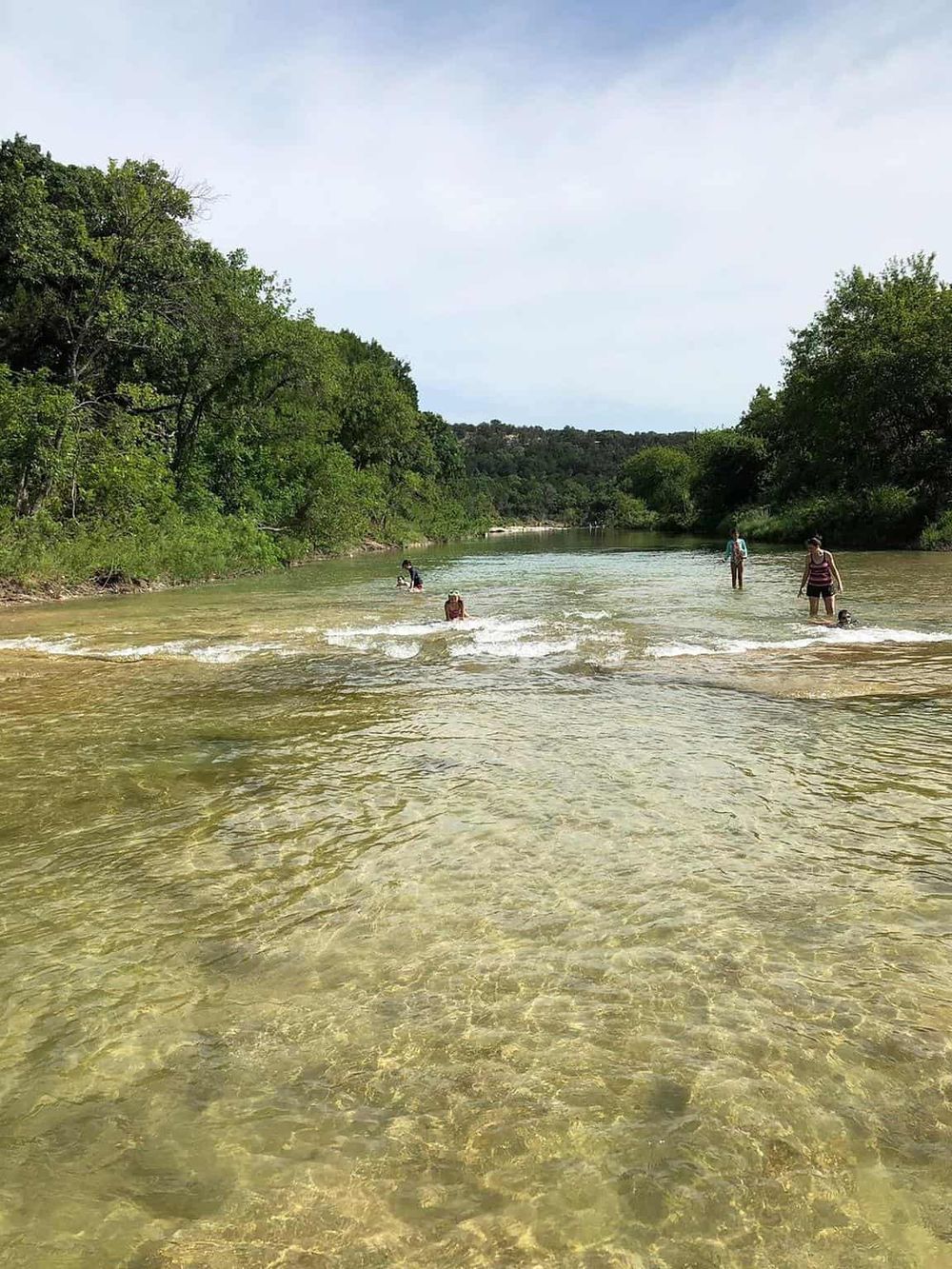 Children playing in the clear river with lush green trees and blue sky in the background.