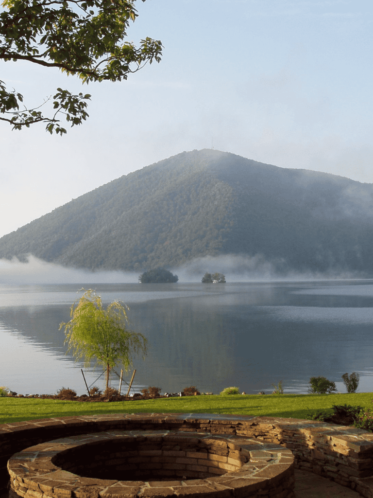 Serene mountain lake scenic view with mist, trees, and outdoor firepit, perfect for nature and adventure seekers.
