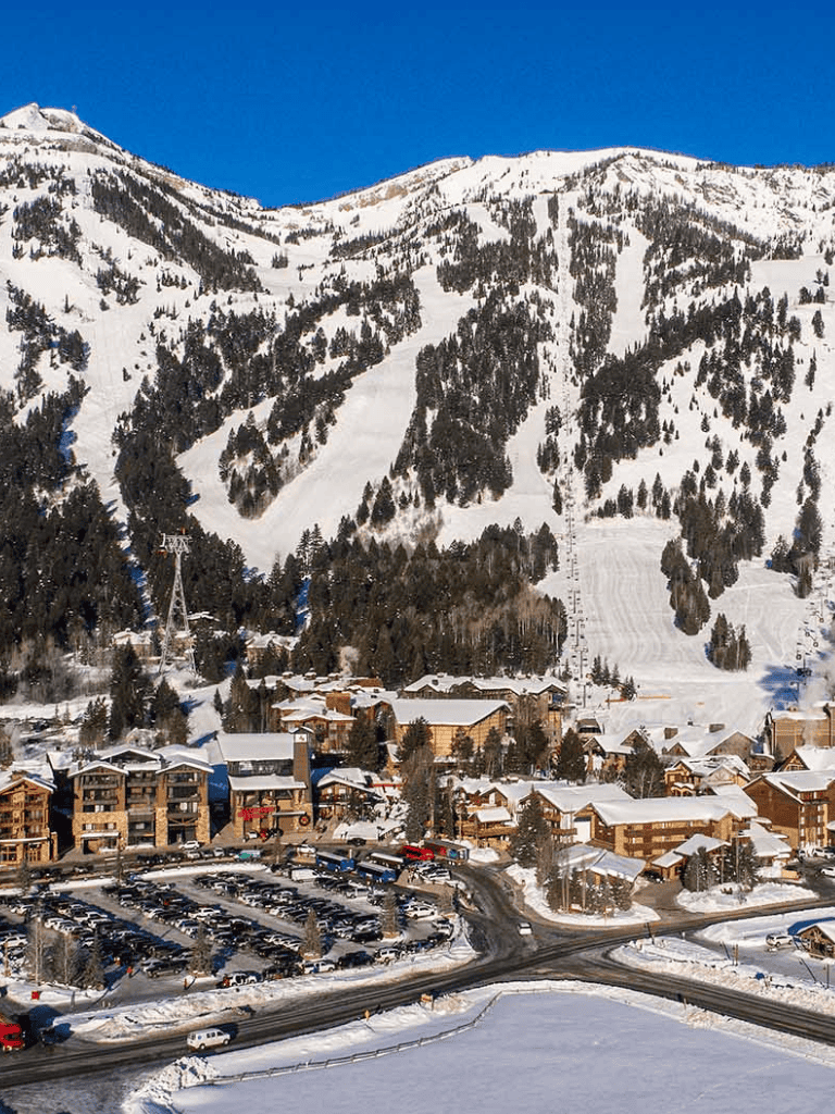 Snow-covered mountain ski resort with alpine village and parking lot at the base, under a clear blue sky.