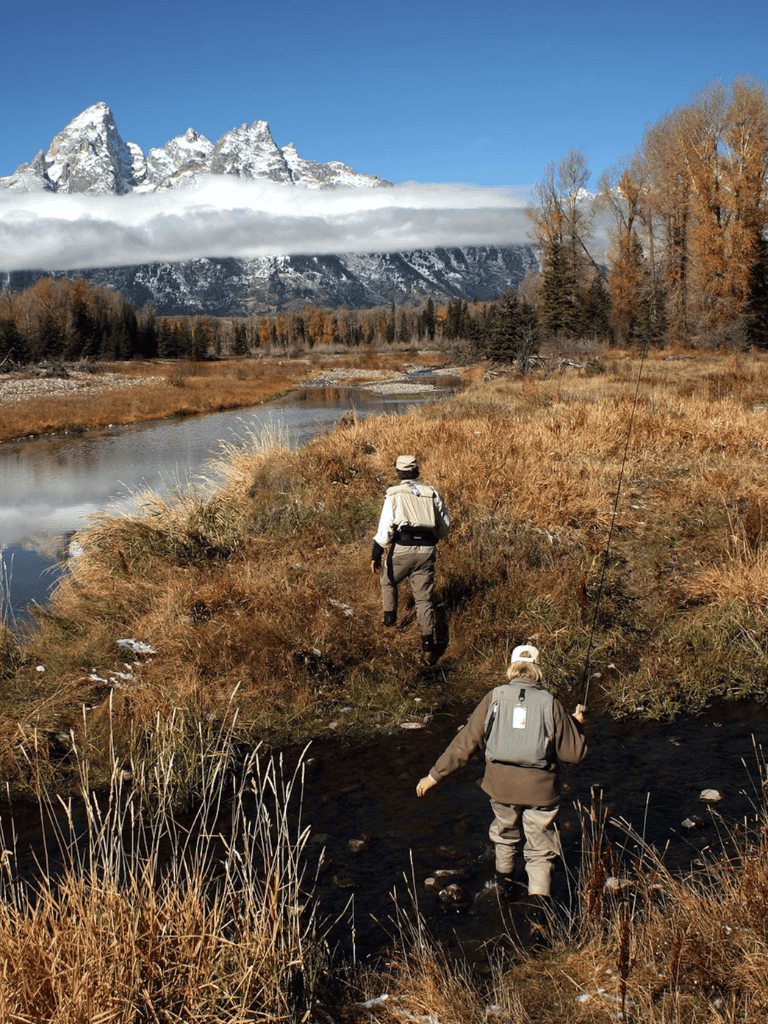 1. Two hikers fishing in scenic mountain river with snowy peaks and autumn trees in the background.