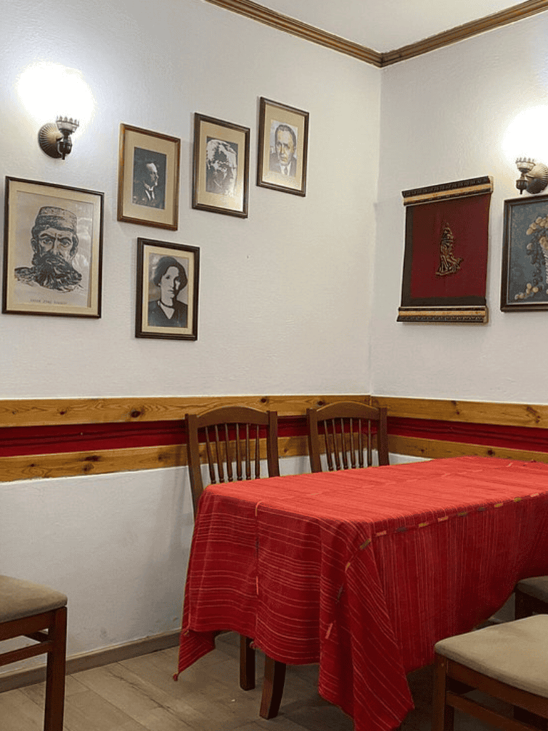 A cozy dining area with framed portraits on white walls and a red tablecloth on the table, featuring warm lighting and classic decor.