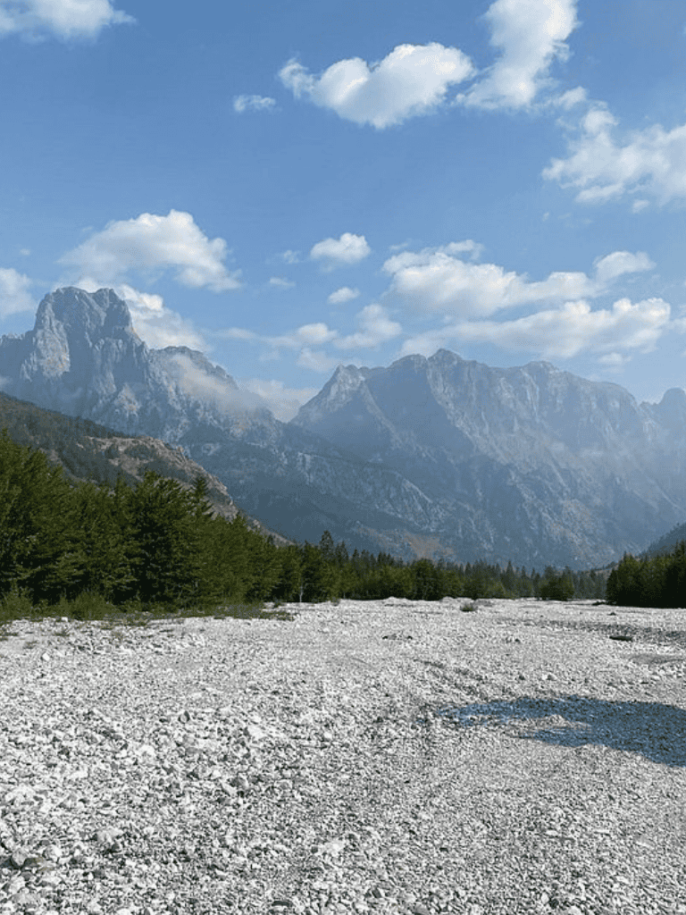 Breathtaking mountain landscape with rocky riverbed, lush green trees, and a bright blue sky with fluffy clouds.