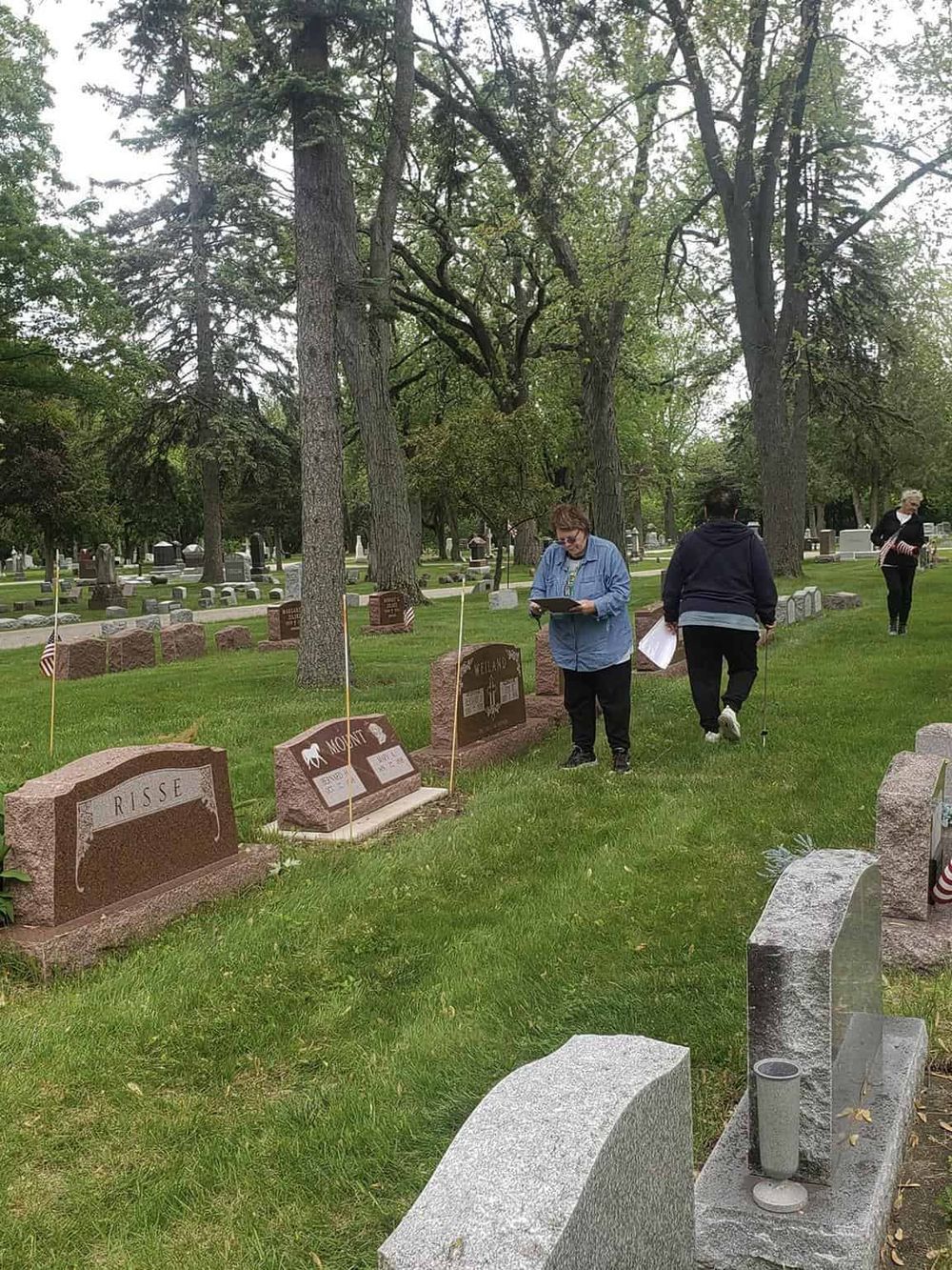 Graveyard with visitors paying respects, surrounded by tall trees and headstones, emphasizing memorial and cemetery exploration.