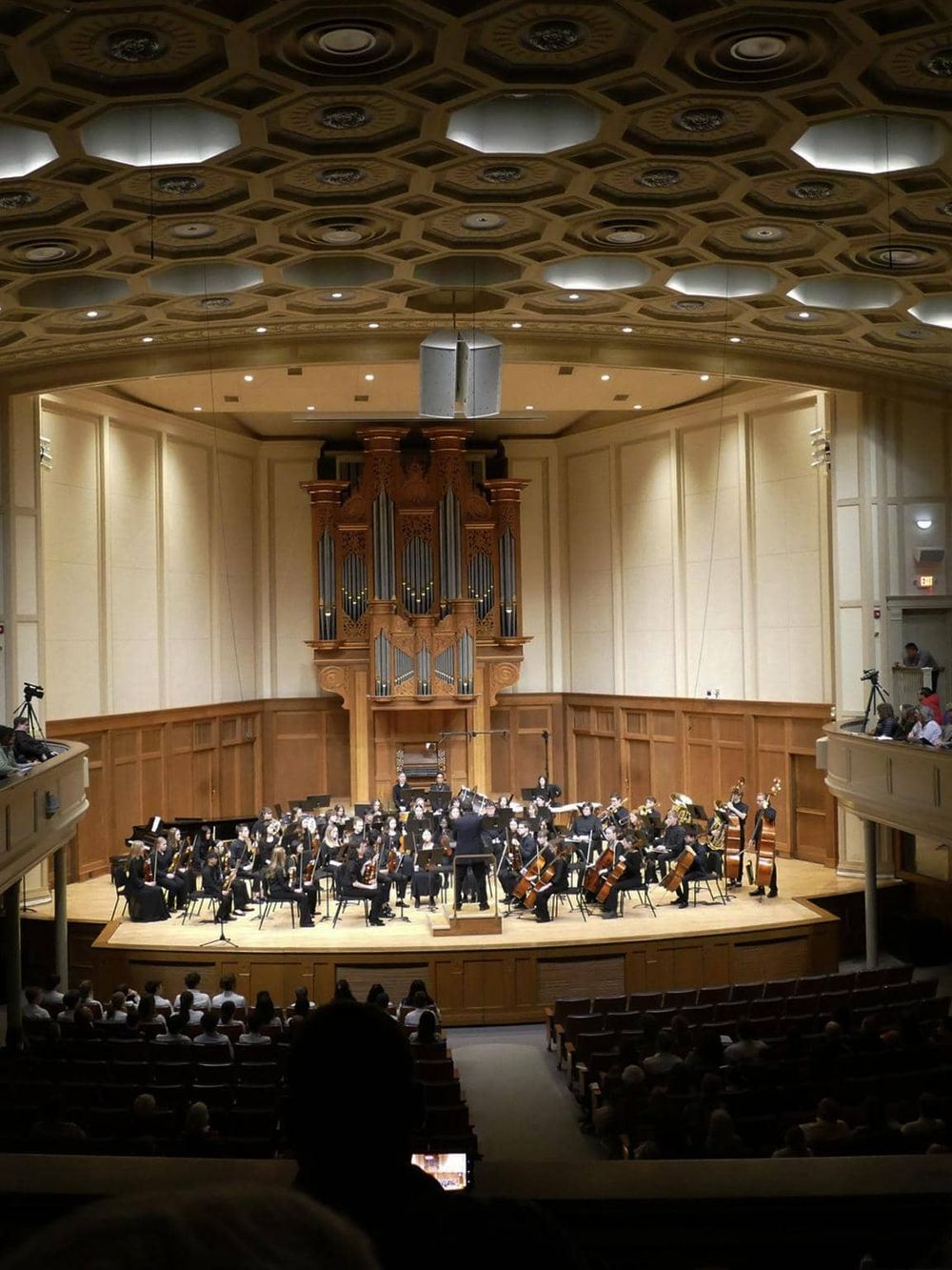 Orchestra performing in a grand concert hall with a wooden stage and ceiling design, elegant acoustics, and audience members watching.