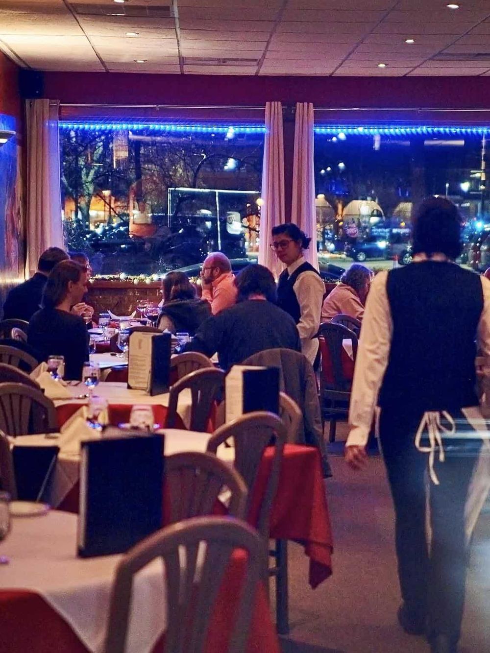 Cozy restaurant dining scene with waitstaff, guests, and large window view of parked cars.