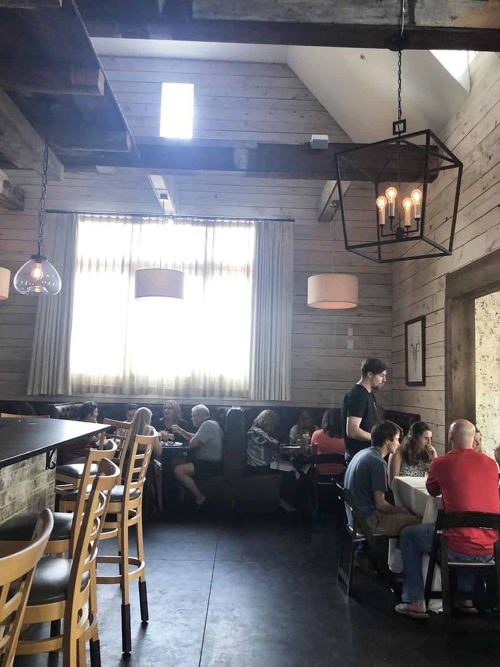Cozy rustic restaurant interior with wooden walls, large window, and warm lighting, perfect for dining and socializing.