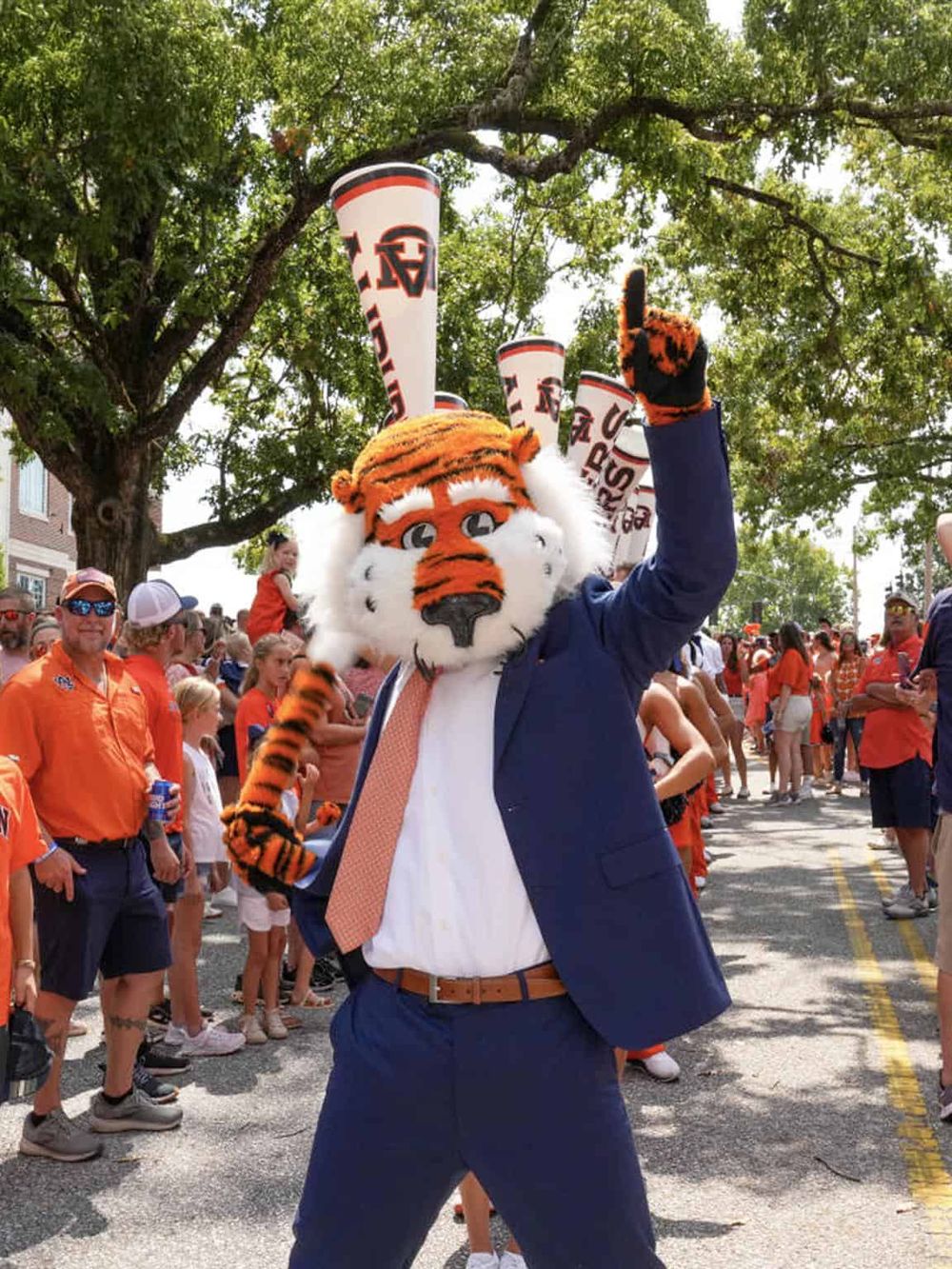 Vibrant image of a university mascot in a suit at a parade, surrounded by students and supporters, under lush trees.