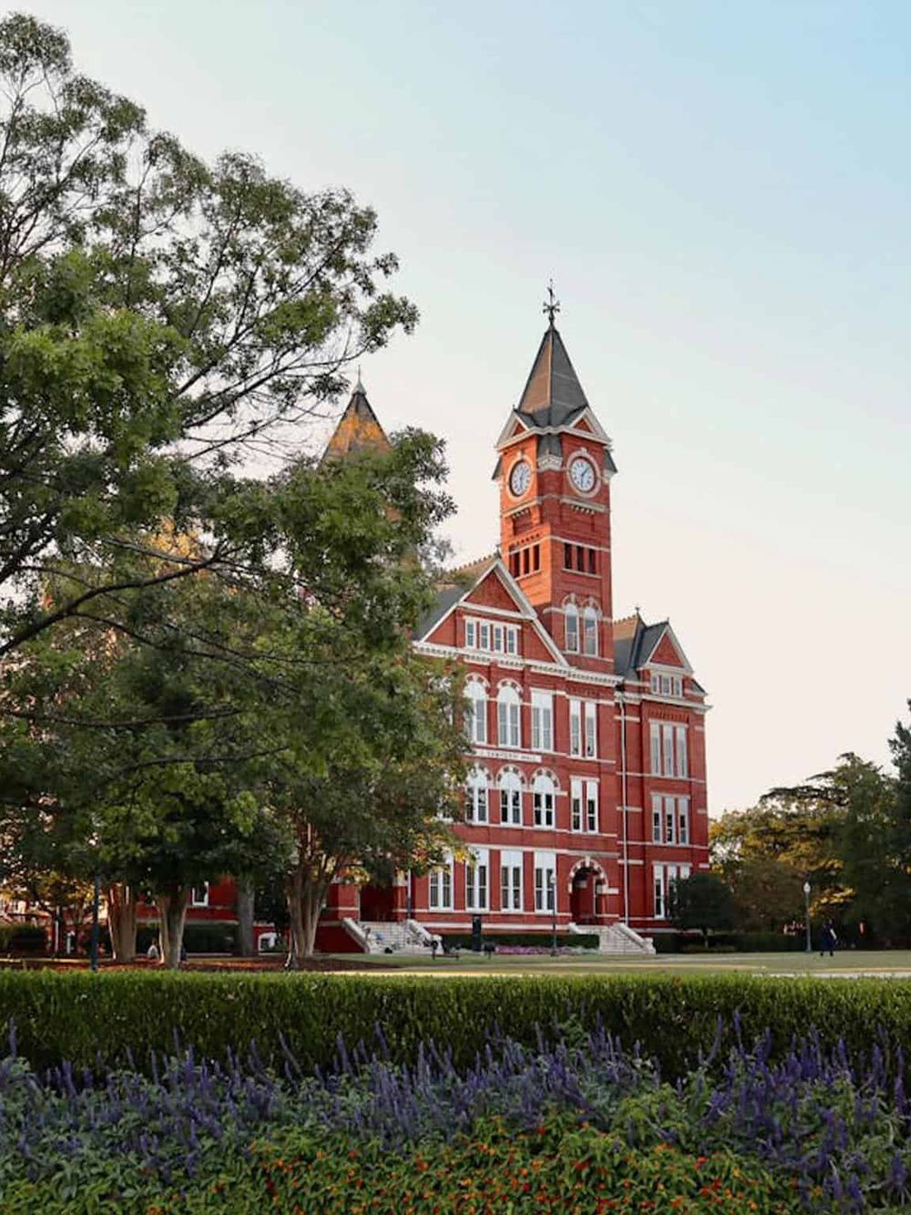 Historic red brick courthouse with clock tower surrounded by greenery and flowers in a scenic park setting.