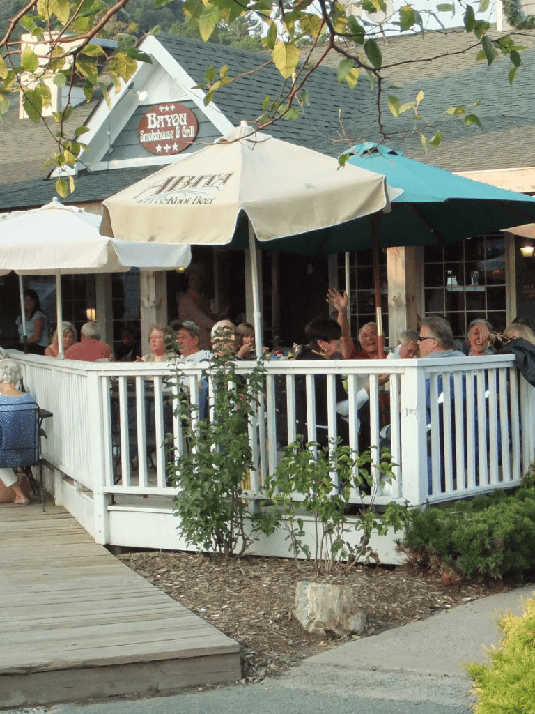 Outdoor restaurant patio with people dining under umbrellas, surrounded by greenery.