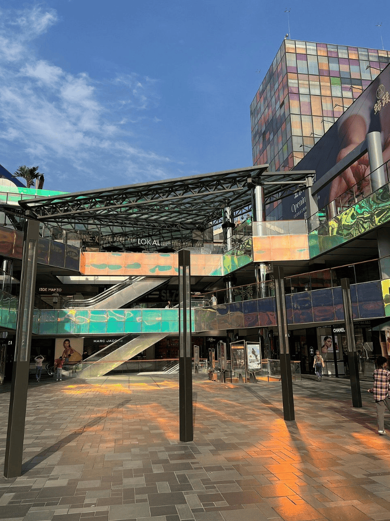 Bright outdoor shopping mall with colorful glass buildings, open walkways, and escalators.