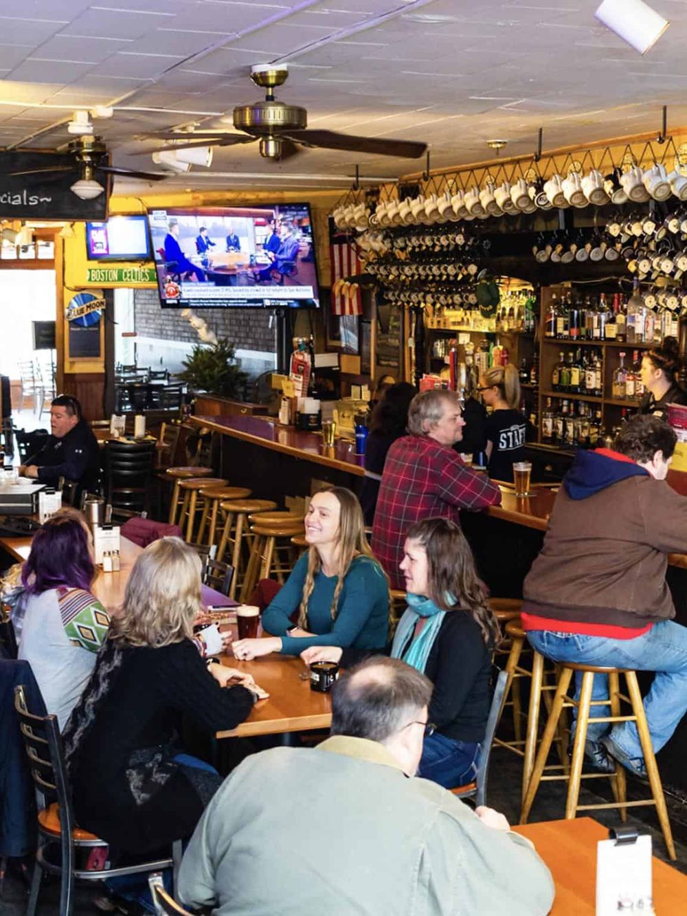 Cozy bar with wall-mounted glasses, patrons enjoying drinks, and a TV displaying a sports event.