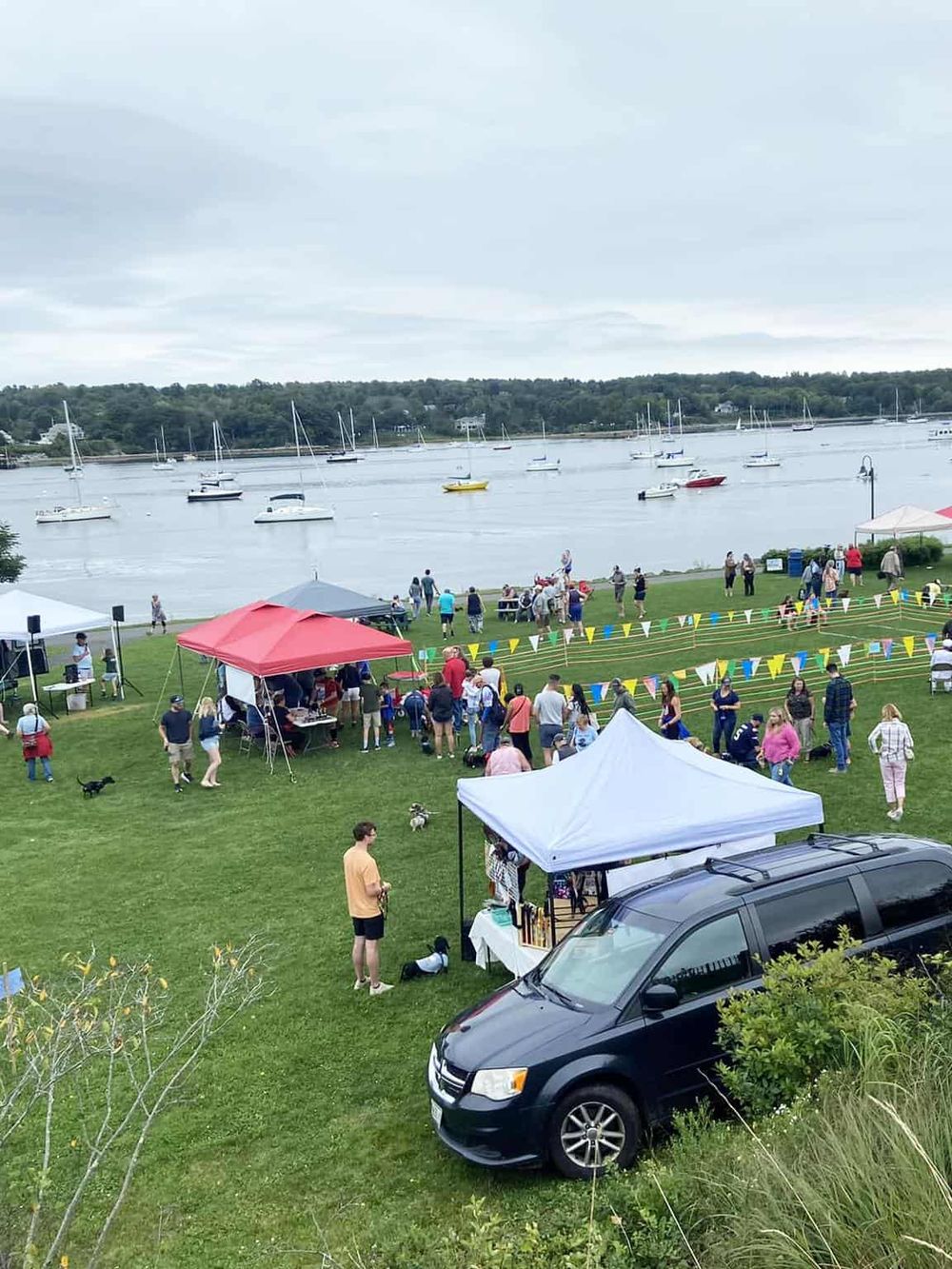 Colorful outdoor event near a waterfront with tents, boats, and people enjoying a community gathering.