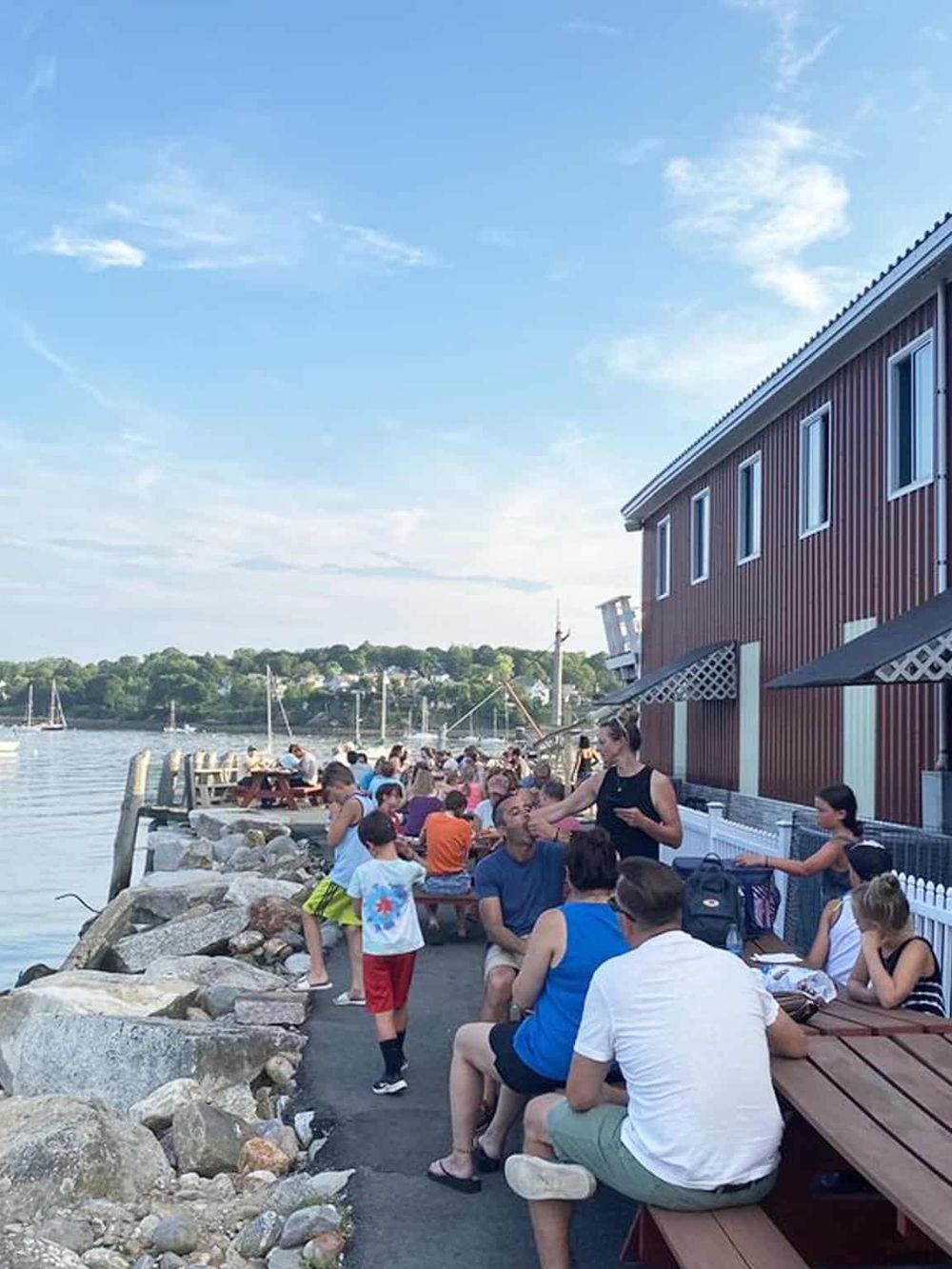 Vibrant waterfront restaurant with people enjoying food and drinks, scenic harbor views, and lively summer atmosphere.