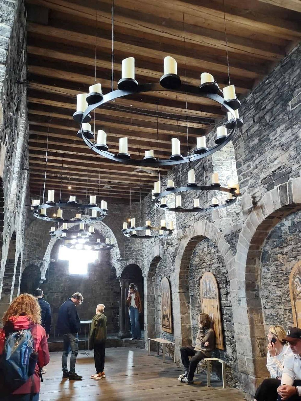 Elegant rustic stone church interior with large chandelier, visitors, and historic artwork, showcasing architecture and atmosphere.