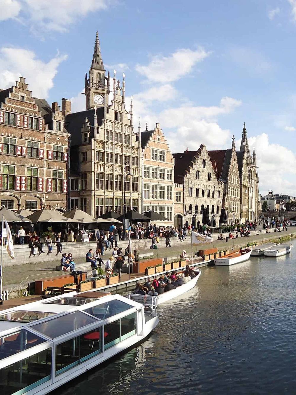 Colorful historic buildings along a canal with boats and people enjoying the scenic waterfront in Belgium.