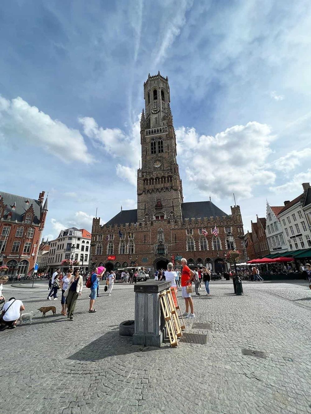 Historic Belfry Tower in Bruges, Belgium, bustling square with visitors and outdoor cafes.