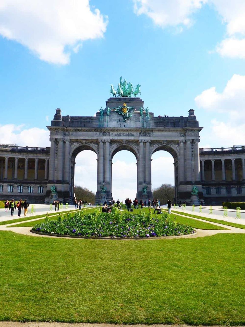 Elegant triumphal arch with a statue of a chariot at the top, set against a bright blue sky, surrounded by green lawns and visitors.