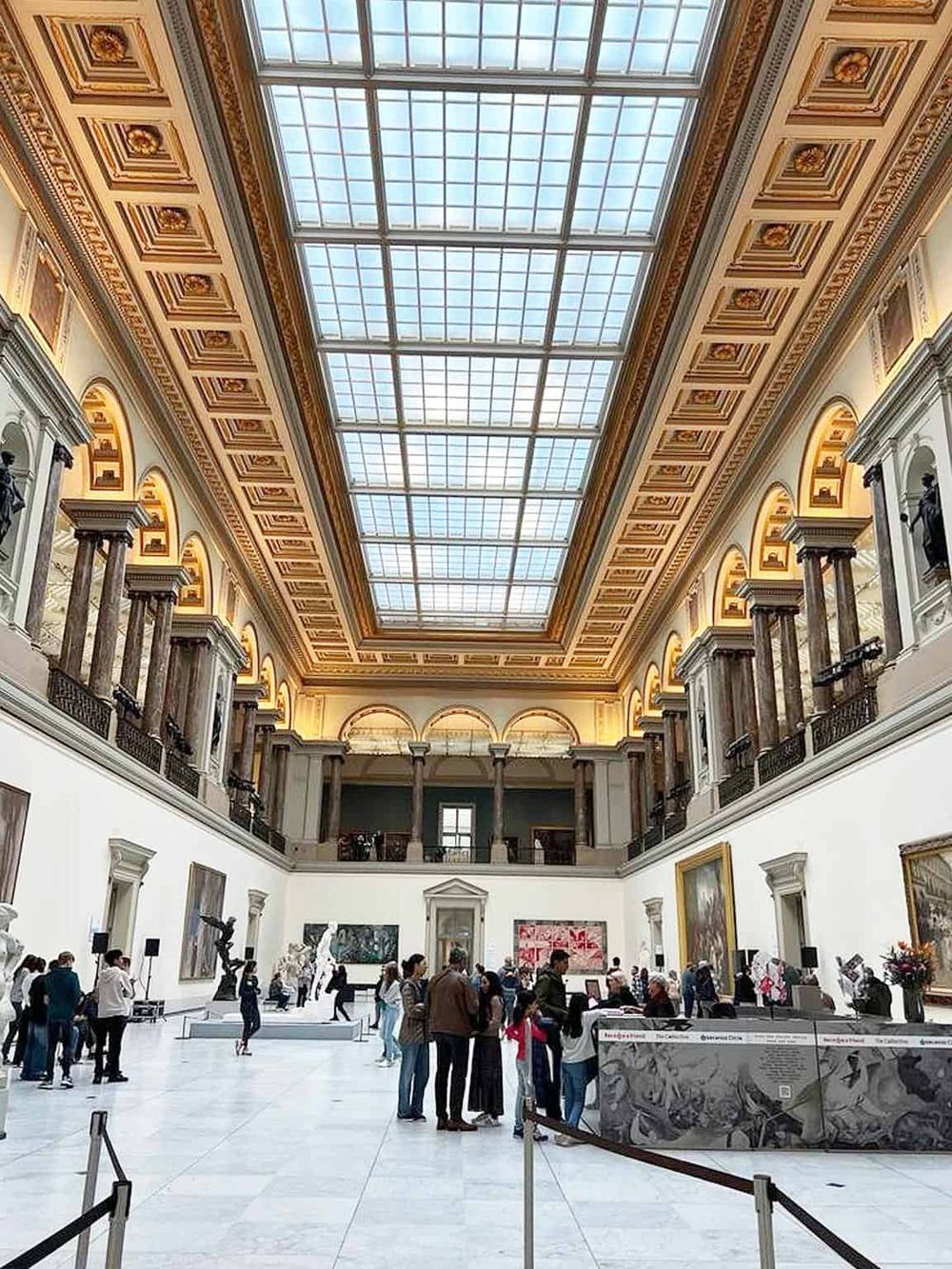 Intricate museum interior with a high glass ceiling, classical architecture, and visitors exploring art exhibits.